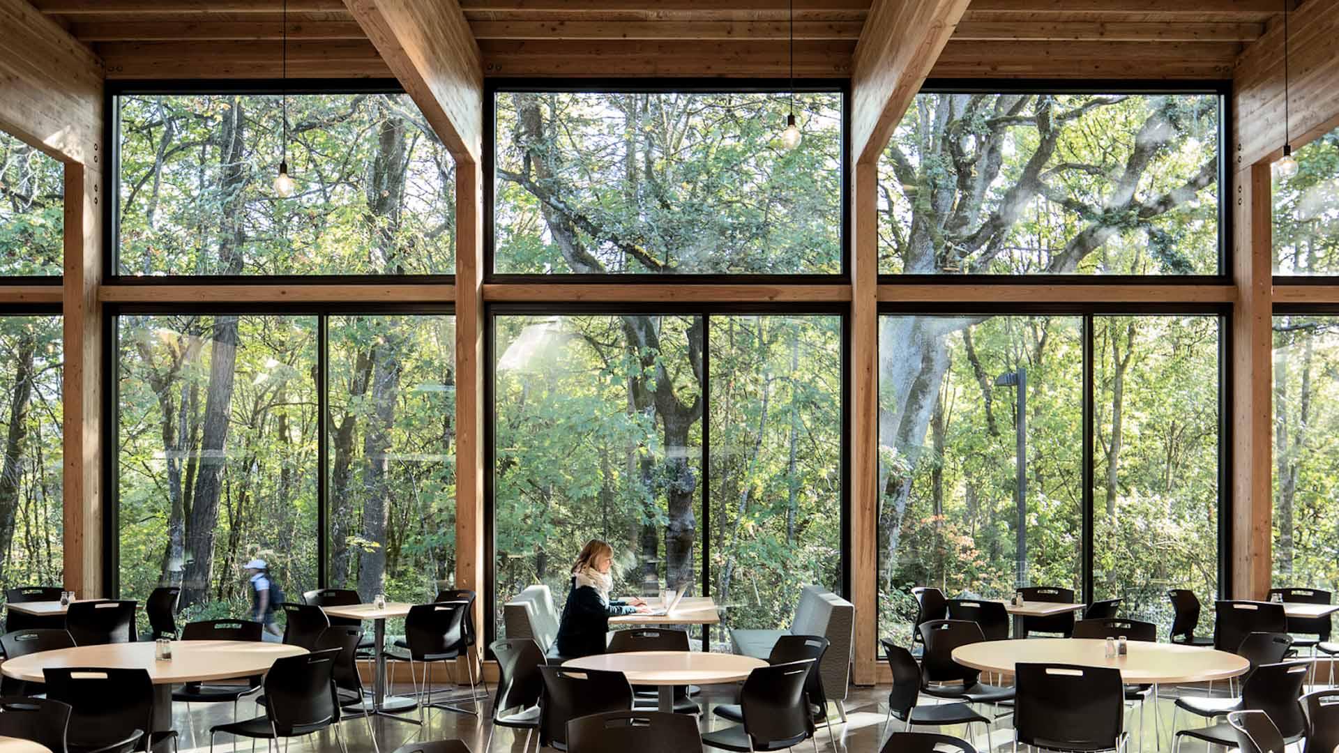 峡谷公地照片——一大片木结构建筑的一大排窗户向外眺望一片树木繁茂的区域. 一位女士坐在前景中的一张桌子旁.